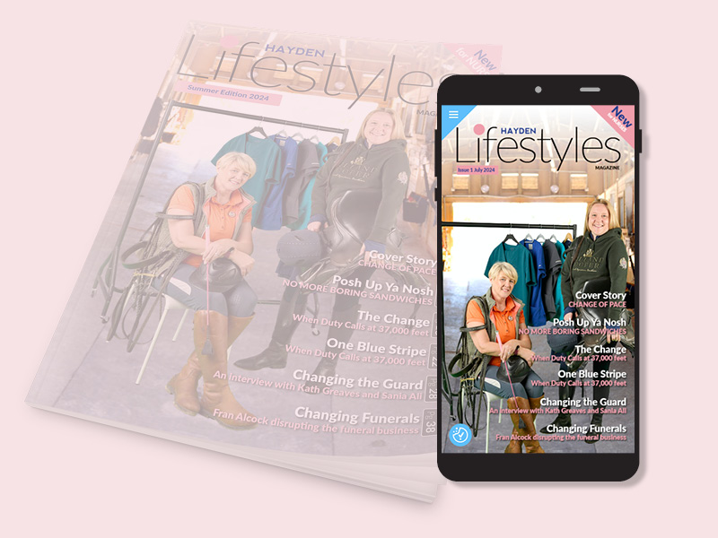 A smartphone displaying the cover of Hayden Lifestyles magazine, featuring two smiling women seated in an equestrian setting. Behind them are jackets on hangers. A printed copy of the magazine is slightly blurred underneath.