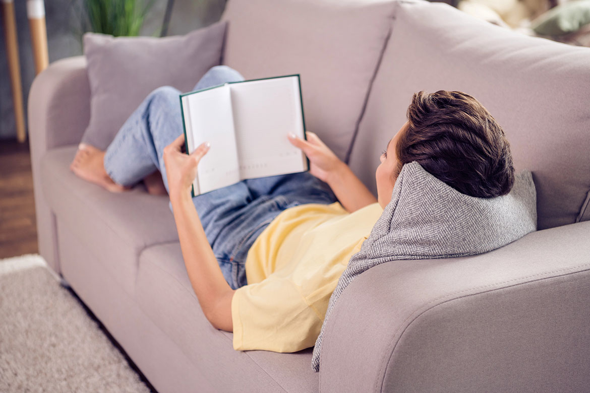 A person in a yellow shirt and blue jeans is lying on a gray couch, resting their head on a pillow, and reading an open book.