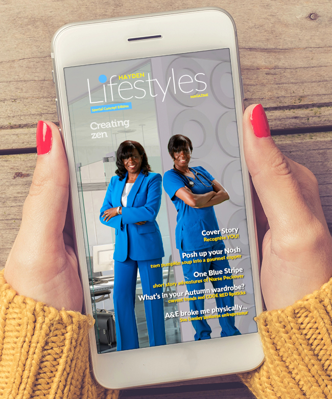 Close-up of a smartphone held in two hands, displaying a lifestyle magazine cover. The cover features a woman portrayed as both an entrepreneur in a blue suit and a nurse in scrubs. The background behind the phone is a weathered wooden table, and the user wears a chunky yellow knit sleeve.