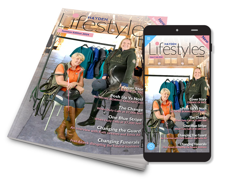 A smartphone displaying the cover of Hayden Lifestyles magazine, featuring two smiling women seated in an equestrian setting. Behind them are jackets on hangers. A printed copy of the magazine is slightly blurred underneath.